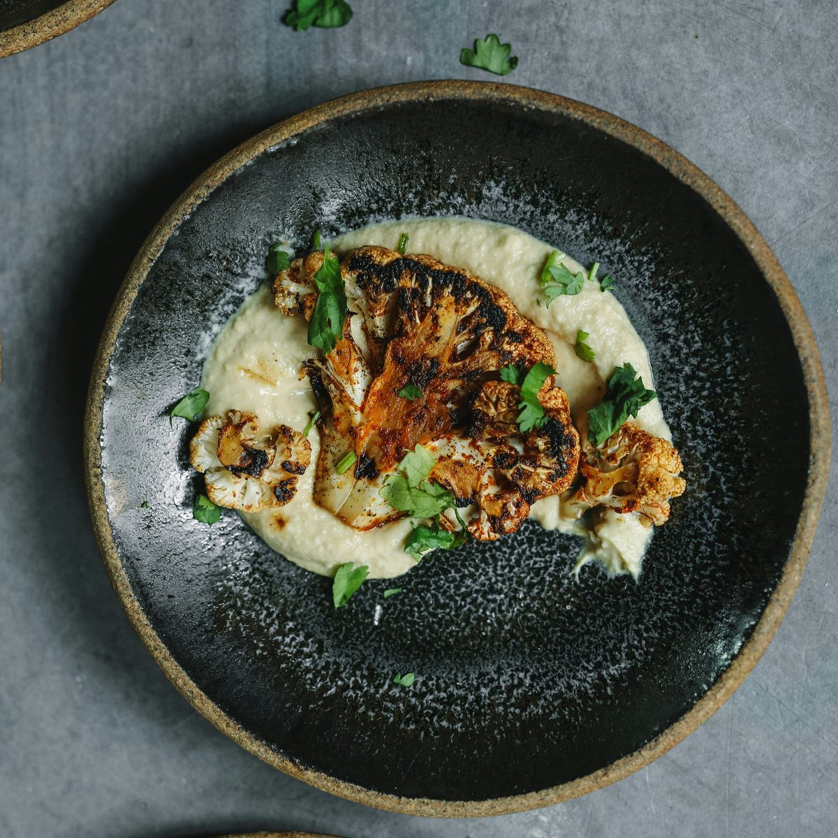 A charred cauliflower steak over whipped tahini with coriander on a dark speckled plate, seen from above