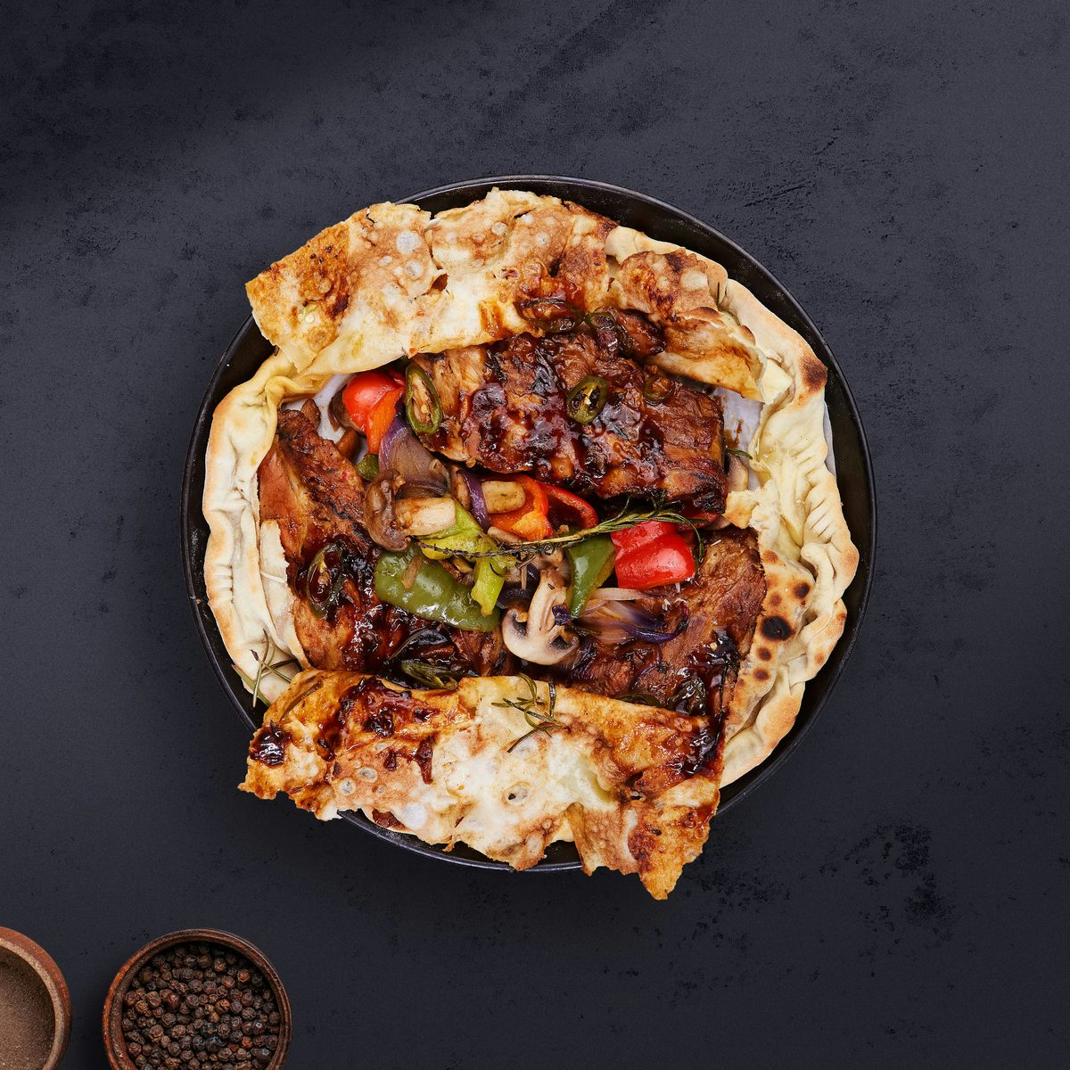 Glazed beef with peppers and mushrooms inside a blistered flatbread parcel on a dark plate, seen from above