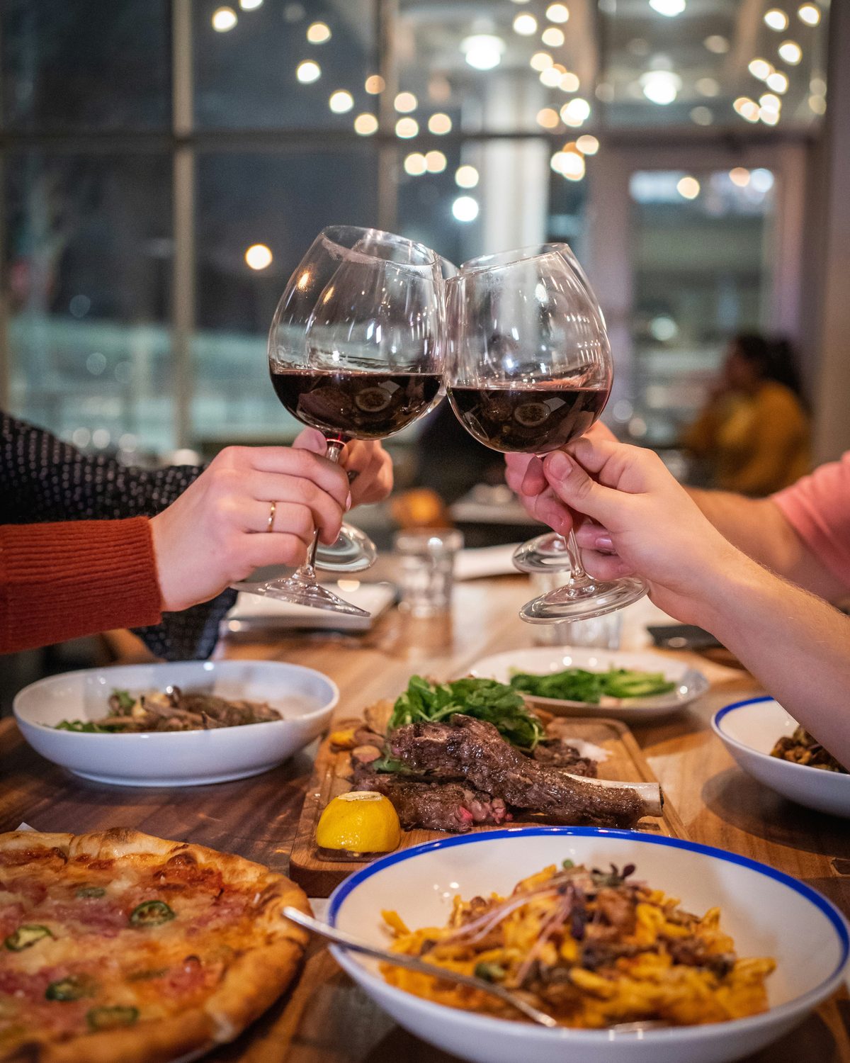 Glasses of red wine raised over a shared table of steak, pasta and pizza